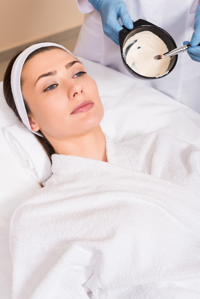 beautician mixing face mask while woman lying on bathrobe and hairband at beauty salon - Photo, Image