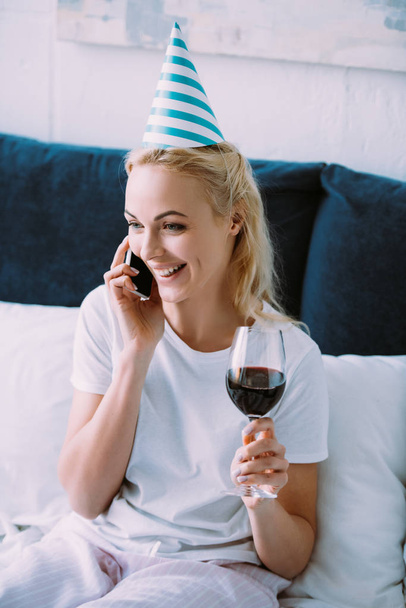 smiling woman in party hat celebrating birthday, holding glass of wine and talking on smartphone in bed at home - Fotografie, Obrázek