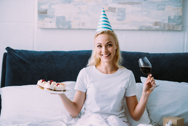 happy woman in party hat holding glass of red wine and cake while celebrating birthday in bed alone - Photo, Image