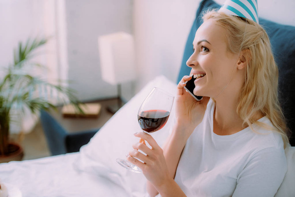 Szczęśliwa Kobieta w Czapka Urodzinowa obchodzi urodziny, trzymając kieliszek wina i mówienia na smartfonie w łóżku w domu - Zdjęcie, obraz