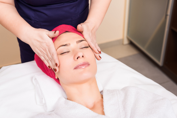 Kosmetyczka robi masaż ręczny twarzy kobieta z czerwony ręcznik na głowę w salonie kosmetycznym - Zdjęcie, obraz