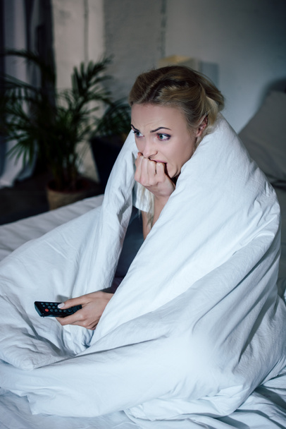 korkmuş kadın battaniye holding uzaktan kumanda, tırnaklarını ısırma ve evde yatakta tv izlerken kaplı - Fotoğraf, Görsel