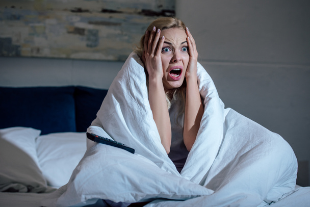 φοβισμένη γυναίκα που καλύπτονται μέσα κουβέρτα με τα χέρια στο κεφάλι ουρλιάζοντας ενώ παρακολουθείτε τηλεόραση στο κρεβάτι στο σπίτι - Φωτογραφία, εικόνα