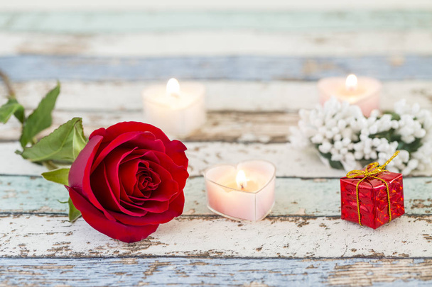 Yksi punainen ruusu, lahjapakkaus, kynttilät ja valkoiset kukat turkoosin pöydällä
 - Valokuva, kuva