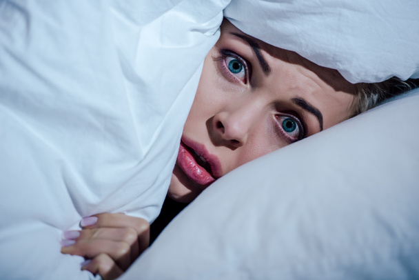 scared woman hiding behind blanket and looking at camera in bedroom at home - Photo, Image