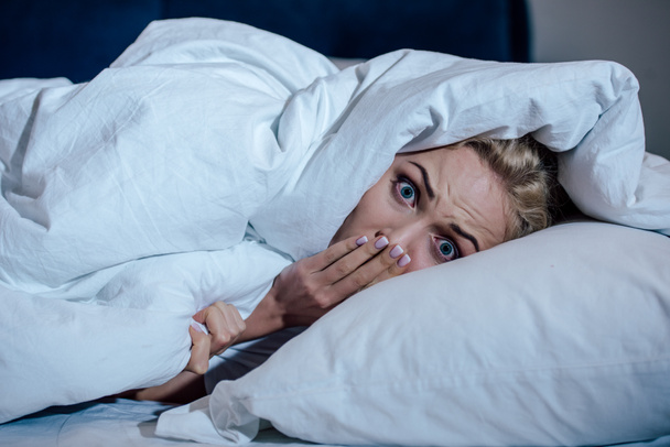 scared woman covering mouth with hand while looking at camera in bed at home - Photo, Image
