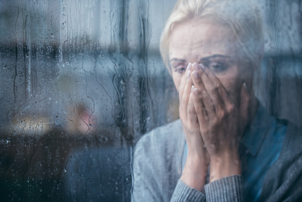 mulher adulta triste chorando e cobrindo rosto com as mãos em casa através da janela com gotas de chuva
 - Foto, Imagem