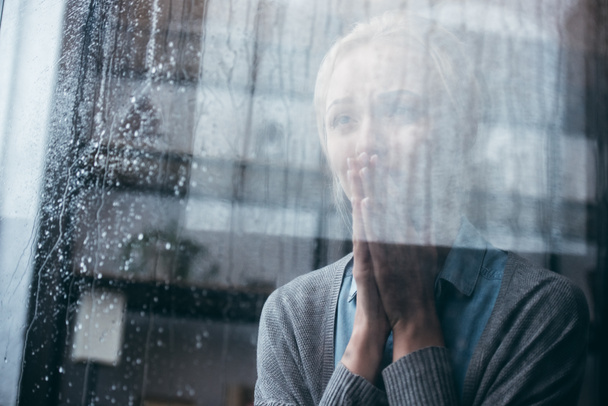 triste mujer adulta con gesto de oración llorando en casa a través de la ventana con gotas de lluvia
 - Foto, Imagen