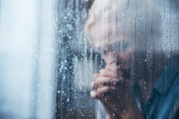 sad adult woman with folded hands at home through window with raindrops - Photo, Image