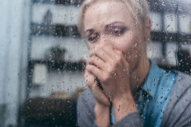 mulher adulta deprimida com as mãos dobradas em casa através da janela com gotas de chuva
 - Foto, Imagem