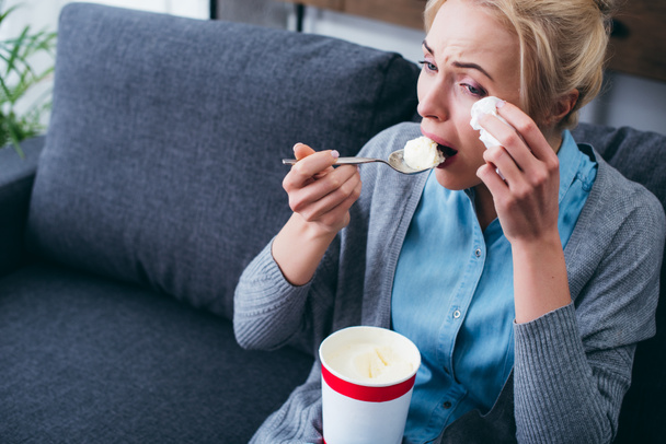 Frau isst Eis und weint, während sie allein zu Hause auf der Couch sitzt - Foto, Bild
