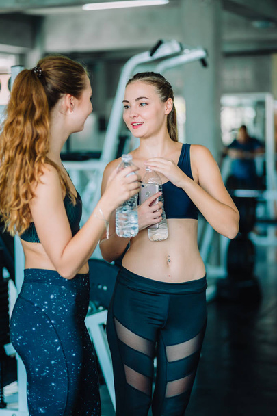 ジムでのトレーニングの後フィットネス服リラックス フィットの美しい若い女性選手。カップルの女性が保持水は瓶の中を立っています。健康、スポーツ、ライフ スタイルのコンセプト. - 写真・画像