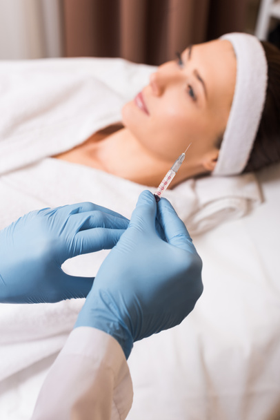 selective focus of syringe in cosmetologist hands in front of woman at beauty salon - Photo, image