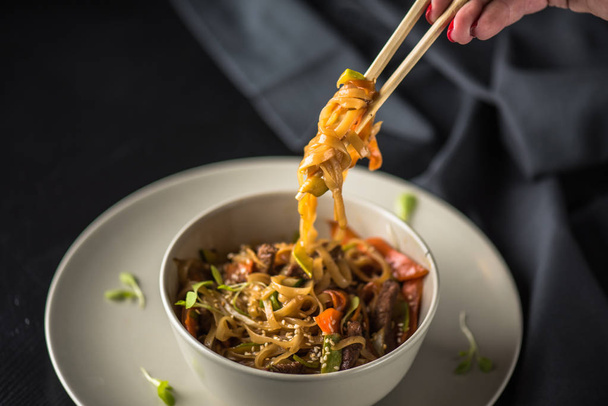 Female hand holding chopstick over bowl of asian seafood noodles on dark background - Фото, изображение