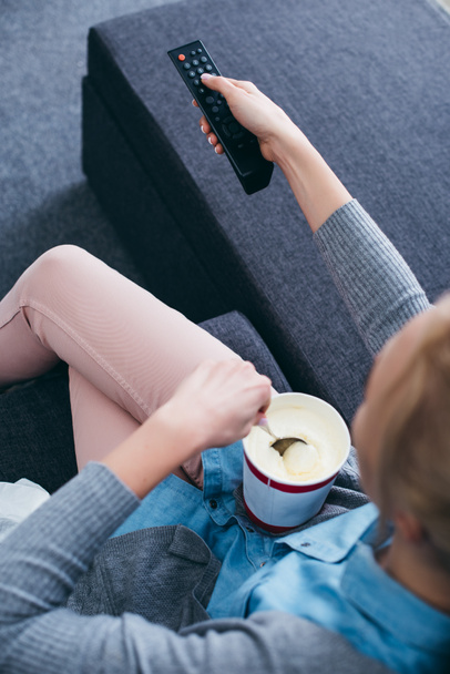 abgeschnittene Ansicht einer Frau, die mit Eis und Fernbedienung auf der Couch sitzt, während sie fernsieht - Foto, Bild