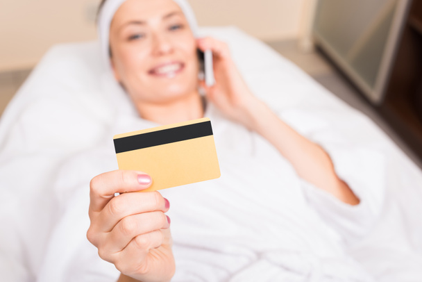 attractive woman lying, talking on smartphone and holding credit card at beauty salon - Zdjęcie, obraz