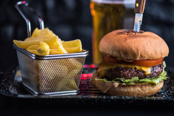 Beef burger served on platter with fried potato wedges on dark background - Фото, изображение
