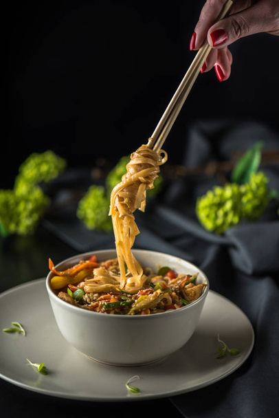 Female hand holding chopstick over bowl of asian seafood noodles on dark background - Fotó, kép