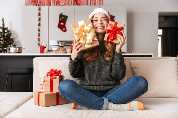Image of cheerful girl 20s sitting on sofa in living room with gift box while celebrating New Year - 写真・画像