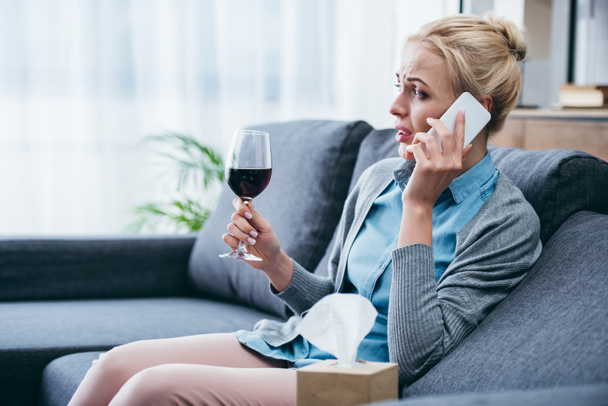 upset woman sitting on couch, holding glass of red wine and talking on smartphone at home - Φωτογραφία, εικόνα