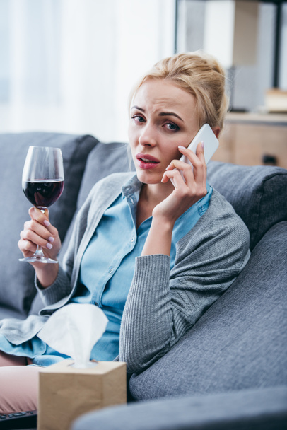 woman sitting on couch, looking at camera, holding glass of red wine and talking on smartphone at home - Photo, Image