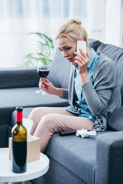 mujer sentada en el sofá, sosteniendo una copa de vino tinto y hablando en el teléfono inteligente en casa
 - Foto, Imagen