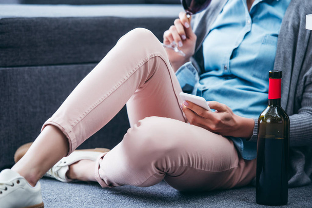 cropped view of woman with bottle of wine sitting and using smartphone at home - Foto, Imagem