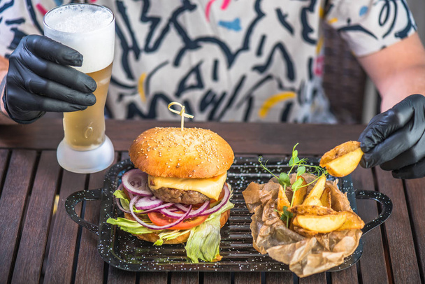 human hands in black gloves with burger, fried potato and beer on table - Фото, изображение