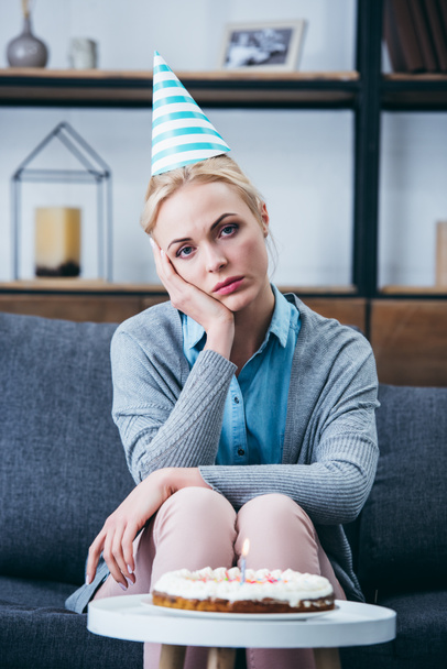 sad woman in party hat propping head with hand and looking at camera while celebrating birthday at home alone - Foto, afbeelding