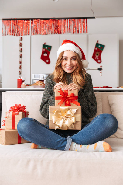 Photo of cheerful woman sitting on sofa in living room with gift boxes while celebrating New Year - Zdjęcie, obraz