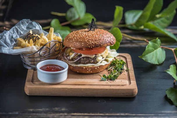 Burger with potato fries served on wooden board on dark background with leaves - Fotó, kép