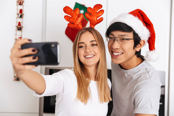 Image of young couple man and woman wearing christmas outfit standing in bright kitchen and taking selfie photo on mobile phone - Fotografie, Obrázek