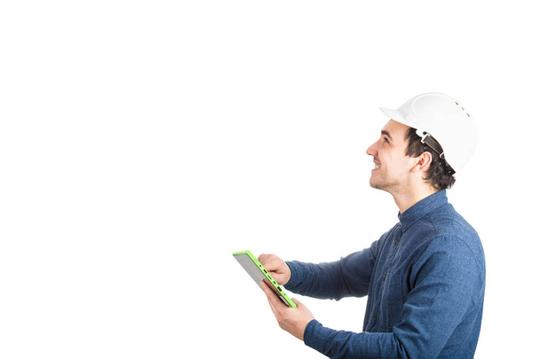 Вид сбоку жизнерадостного молодого человека инженера в защитном шлеме с планшетным компьютерным гаджетом, смотрящим на изолированном белом фоне с копировальным пространством
. - Фото, изображение
