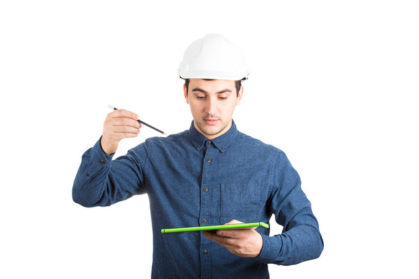 Creativo giovane ingegnere indossando casco protettivo utilizzando un computer tablet e tenendo una matita cercando di disegnare un immaginario schizzo piano 3d isolato su sfondo bianco
. - Foto, immagini
