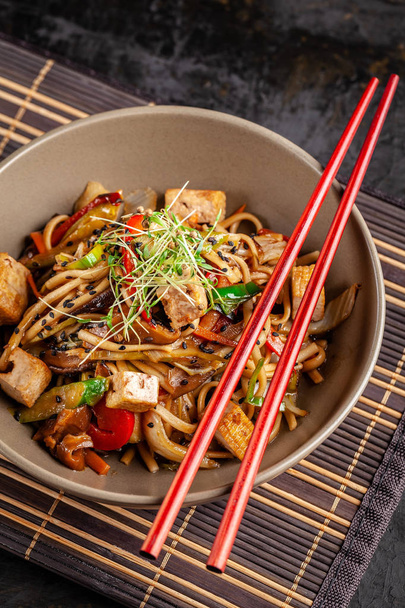 The concept of Japanese cuisine. Chinese noodles with chicken, grilled vegetables, and tofu in unagi sauce. Serving Asian dishes in the restaurant in a plate on a bamboo mat. Top view, copy space - Zdjęcie, obraz