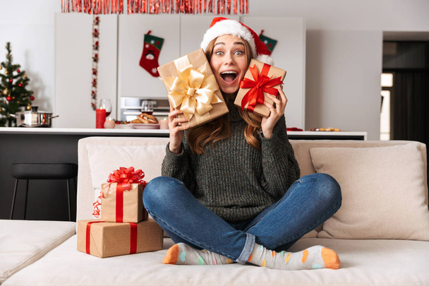 Photo of excited woman sitting on sofa in living room with gift boxes while celebrating New Year - Zdjęcie, obraz