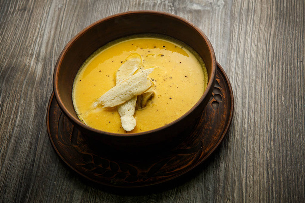 Желтый кремовый суп, украшенный грибами и двумя ломтиками сушеного хлеба, подается в глубокой глиняной тарелке на деревянном столе
 - Фото, изображение