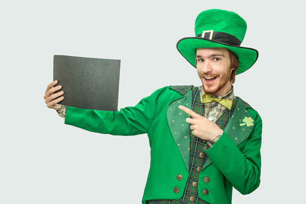 Happy young man in green suit holding black piece of paper. He wears saint Patricks suit. Isolated on grey background. - Foto, Imagen