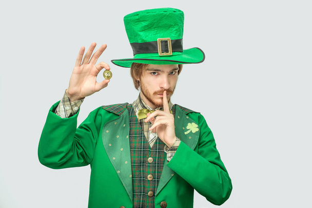 Serious and concentrated young man holding golden coin 100 and look on camera. He hold finger close to mouth. Guy wears saint Patricks suit. Isolated on grey background. - Фото, зображення