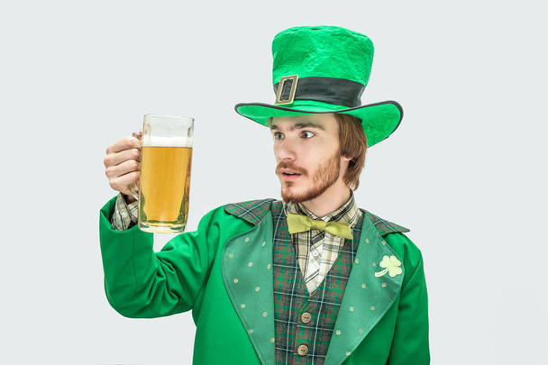 Joven sorprendido en traje verde de San Patricio sosteniendo taza de cerveza y mírala. Está asombrado. Aislado sobre fondo gris
. - Foto, imagen
