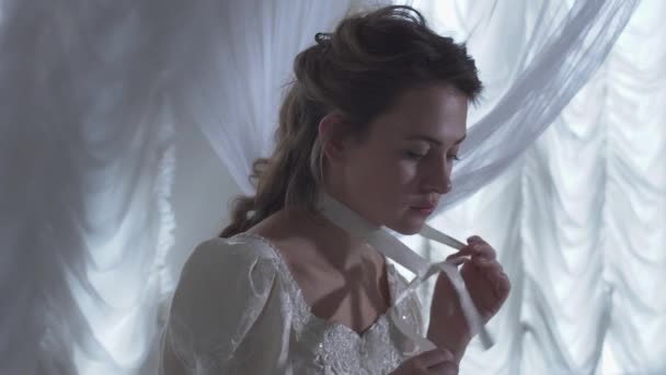 Young beautiful woman sitting arround white net curtains and unties a ribbon on her neck - Imágenes, Vídeo