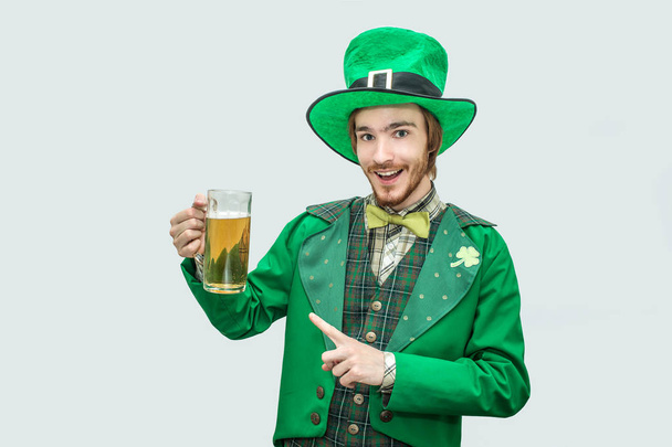 Happy cheerful young man in green saint patick suit holding mug of beer and look on camera. He point on it and smile. Isolated on grey background. - Foto, immagini