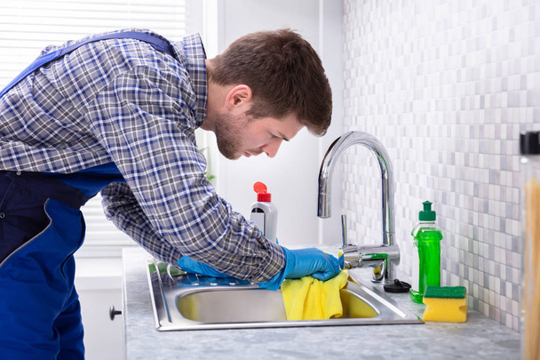 Young Male Janitor Cleaning Kitchen Sink With Napkin And Bottle - Foto, Imagen