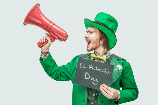 Loud young man in green suit holding dark plate with written words St. Patricks day. Also he speak into red loudspeaker. Isolated on grey background. - Valokuva, kuva