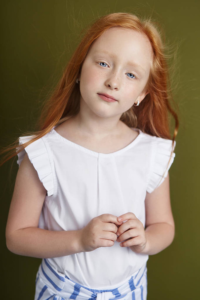 Beautiful red-haired girl with long hair and beautiful big blue eyes. Redhead Girl child in summer clothes posing on the background of olive color. Norwegian girl with bright red hair - Fotó, kép