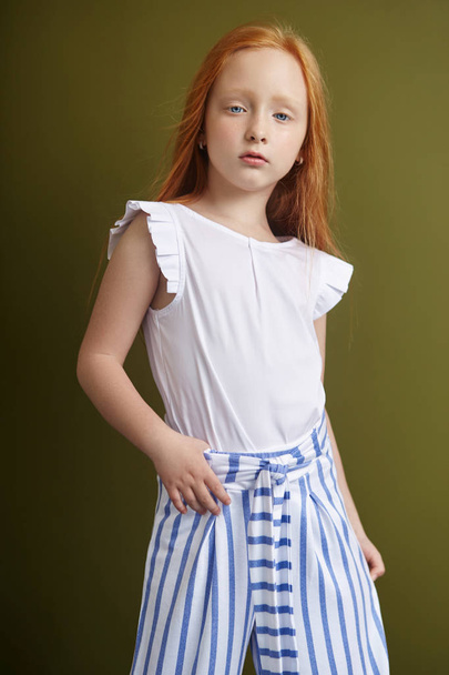 Beautiful red-haired girl with long hair and beautiful big blue eyes. Redhead Girl child in summer clothes posing on the background of olive color. Norwegian girl with bright red hair - Foto, Imagem