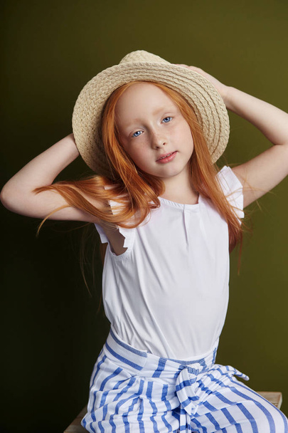 Beautiful red-haired girl with long hair and beautiful big blue eyes. Redhead Girl child in summer clothes posing on the background of olive color. Norwegian girl with bright red hair - Photo, image