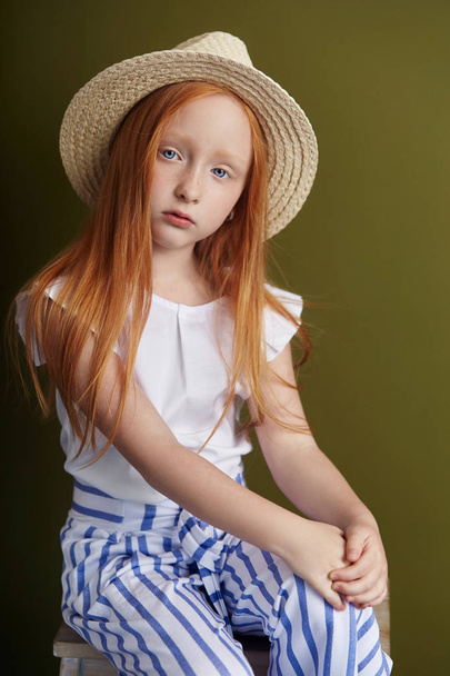 Beautiful red-haired girl with long hair and beautiful big blue eyes. Redhead Girl child in summer clothes posing on the background of olive color. Norwegian girl with bright red hair - Photo, Image