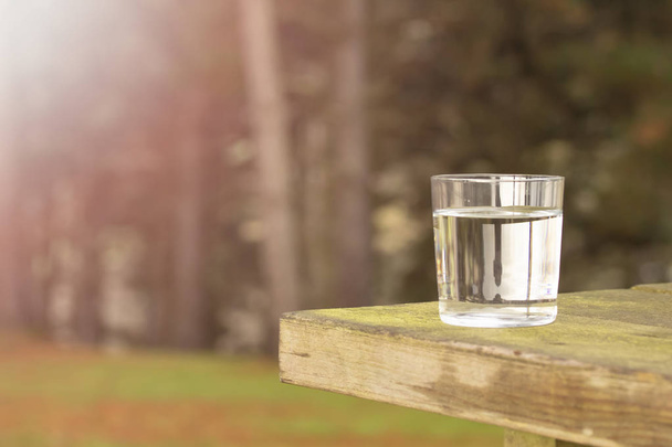 Clear water in a transparent glass on an old wooden table against the background of coniferous forest. Healthy food and ecologically pure natural water from a spring - Photo, Image