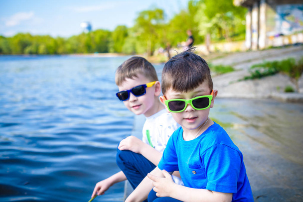 野外自然の中で遊ぶ子供たち: 暖かい夏や春の日にきれいな水で砂に触れる湖または川の海岸に座っています。兄弟の子供男の子一緒に楽しんでいます。友情の概念. - 写真・画像